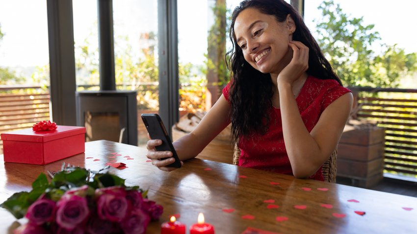 Eine Frau sitzt an einem Holztisch mit dem Smartphone in de rHand. Sie lächelt. Vor ihr liegen Blumen und ein Geschenk.