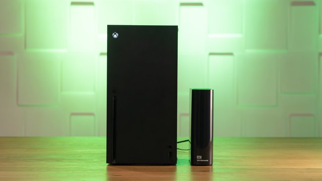 Eine Xbox Series X und eine externe Festplatte von Western Digital stehen auf einem Tisch vor einer grün ausgeleuchteten Wand.