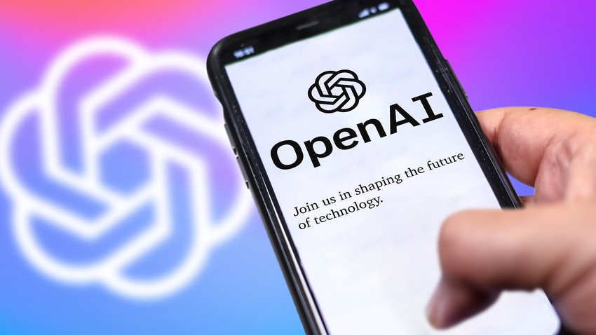 Eine Hand hält ein Smartphone mit geöffnetem ChatGPT. Im Hintergrund ist das Logo von OpenAI zu sehen.