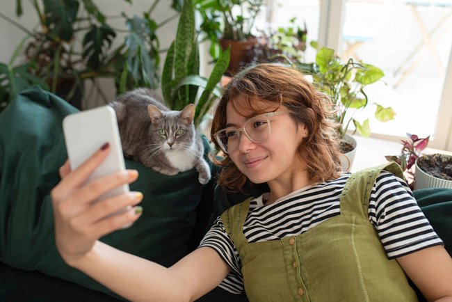 Eine weiblich gelesene Person sitzt auf einem Sofa mit ihrer Katze und macht ein Selfie.