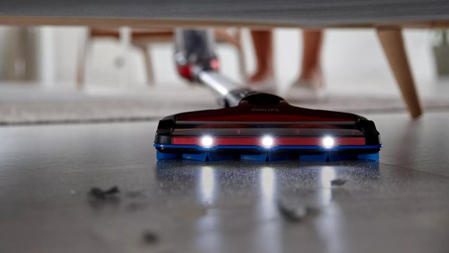 Ein roter Akku-Staubsauger mit LED-Lämpchen saugt einen staubigen Boden. Im Hintergrund sieht man Möbel und die Person, die den Staubsauger hält.