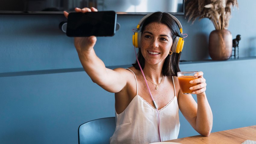 Eine weiblich gelesene Person sitzt an einem Tisch, trägt Kopfhörer und hält mit einer Hand ein Glas mit Saft. In der anderen Hand hält sie ihr Handy für ein Selfie.