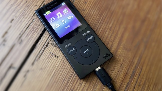 Ein MP3-Player, Modell Sony NW-E393, liegt auf einer Holzdiele. Er ist per USB-Kabel zum Aufladen angeschlossen.