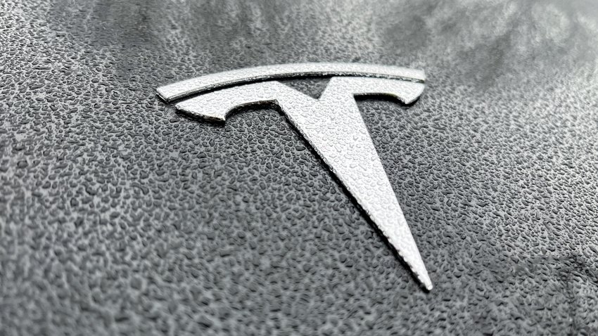 Das silberfarbene Logo des Autoherstellers Tesla auf einer nassen Motorhaube.