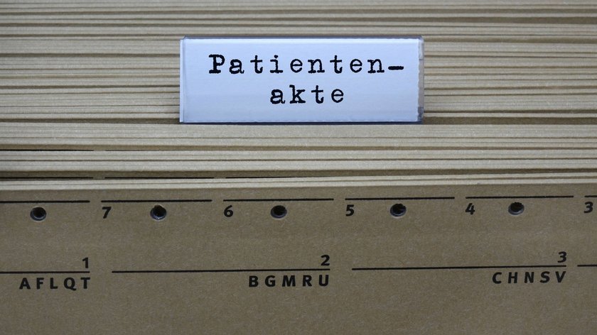 Eine Reihe von braunen Hängeregistermappen mit einem weissen Schild darüber, auf dem "Patientenakte" in schwarzer Schrift steht.