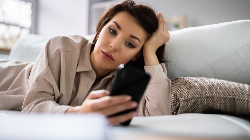 Eine Frau liegt auf dem Bett und schaut traurig auf ihr Handy.