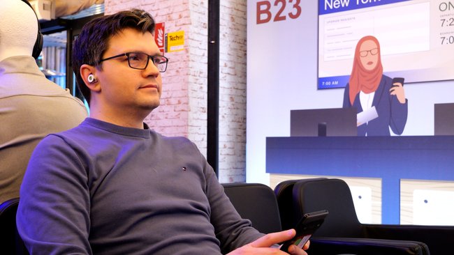 Frank sitzt in einem nachgebauten Flughafen-Set. Ein Smartphone in der Hand, Auracast-In-Ears in den Ohren.