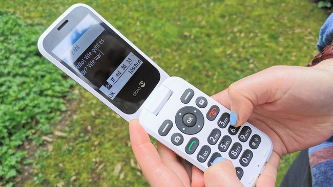 In das aufgeklappte Handy der Marke Doro wird eine SMS getippt, im hintergrund eine grüne Wiese.