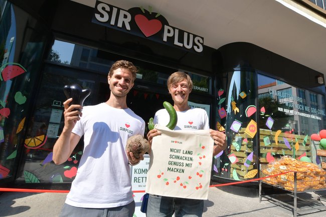 Zwei Männer stehen vor einem Laden und halten Gemüse in die Höhe. Bei dem Laden handelt es sich um SirPlus.