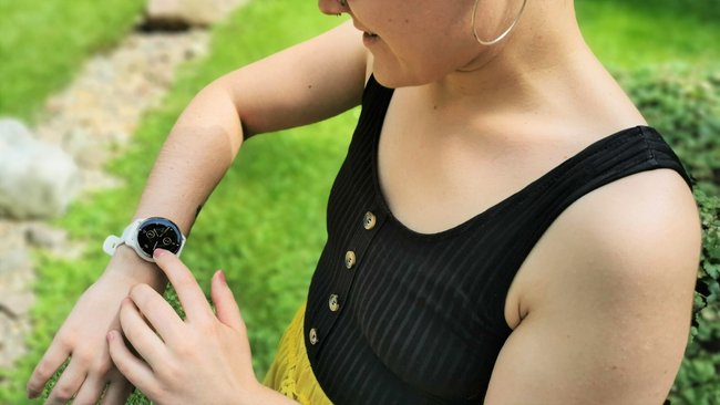 Im Vordergrund ist eine Frau mit schwarzem Oberteil zu sehen. Sie hat den Arm angewinkelt und schaut auf ihre Smartwatch Venu 2 Plus von Garmin. Im Hintergrund ist ein Garten zu sehen.