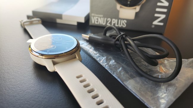 Die weiße Smartwatch Garmin Venu 2 Plus liegt auf einer schwarzen Oberfläche, daneben das Ladekabel mit Plastikhülle. Im Hintergrund ist der Karton zu sehen und die Gebrauchsanweisung.