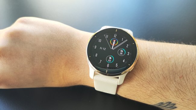 Im Vordergrund sieht man das Handgelenk, an dem die Smartwatch Venu 2 Plus angebracht ist. Das Display zeigt die Uhrzeit und Schrittzahl an. Im Hintergrund sieht man eine schwarze Oberfläche.