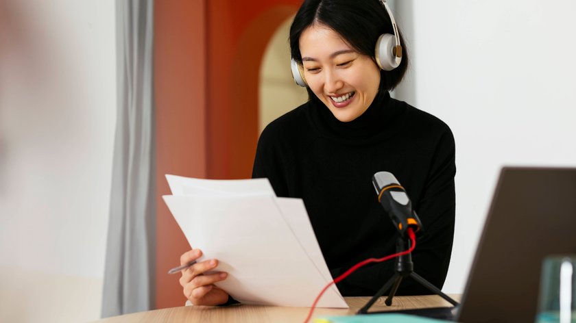 Eine Frau sitzt vor einem Mikrophone, hat Kopfhörer auf und liest sich Notizen durch.