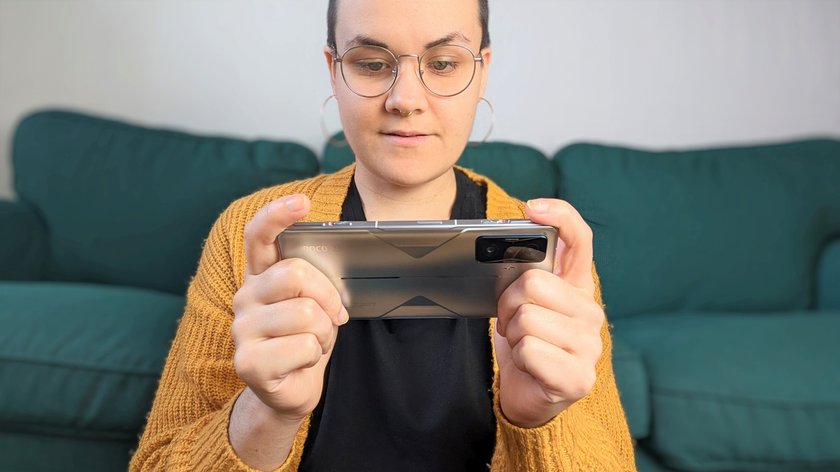 Eine Frau hält das Smartphone Poco F4 GT vor sich in ihren Händen, im Hintergrund steht ein grünes Sofa.