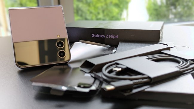 Auf einem schwarzen Tisch liegen das USB-C-Kabel, die Folie und der Karton neben dem dazugehörigen Smartphone. Das Modell ist das Samsung Galaxy Z Flip4.