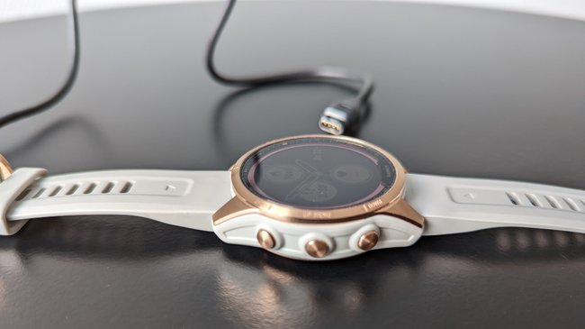 Auf einer schwarzen Tischplatte liegt die Smartwatch Garmin Fenix 7S, dahinter ist das Ladekabel.