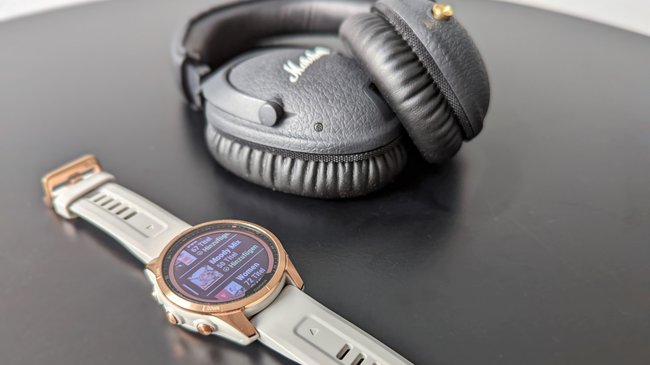 Auf einer schwarzen Tischplatte liegt die Smartwatch Garmin Fenix 7S mit geöffnetem Musikmenü- Im Hintergrund liegt ein Kopfhörer.