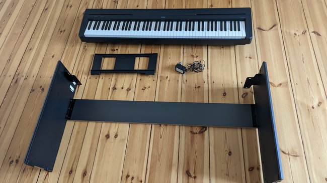 Auf einem Dielenboden liegt ein Digitalpiano mit Notenhalter, Kabel und Ständer.