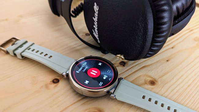 Auf einem Holzbrett liegt die Huawei Watch GT 4, daneben Kopfhörer der Marke Marshall.
