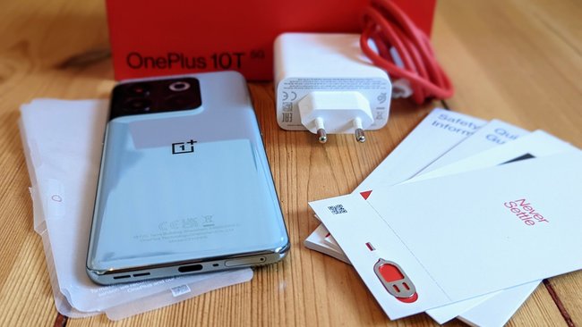 Auf einem Dielenboden liegt der komplette Lieferumfang zum Smartphone OnePlus 10T.