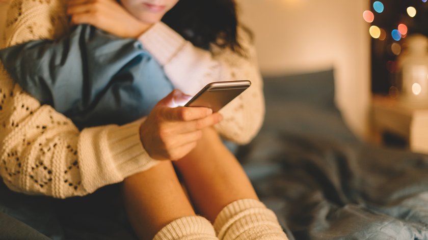 Eine Frau sitzt auf einem Bett und hält ein Smartphone in der Hand.