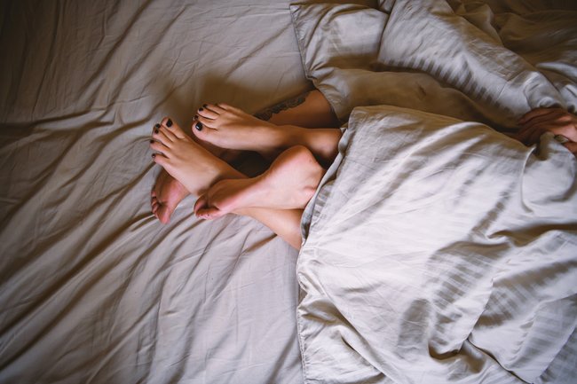 Zwei Menschen lieben in einem Bett, man sieht nur die Füße der zwei unter der Bettdecke.