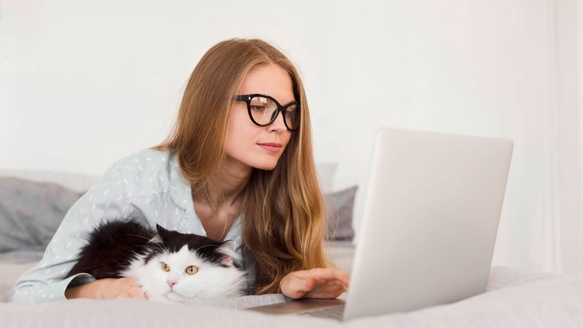 Eine Frau trägt eine Brille und sitt vor ihrem Laptop. Neben ihr liegt ihre Katze.