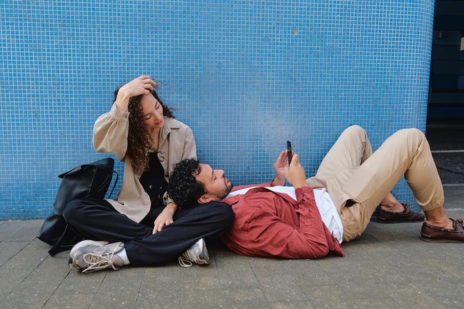 Eine Frau und ein Mann sitzen auf dem Fußweg, er ist an ihr Bein gelehnt und schaut auf sein Smartphone.