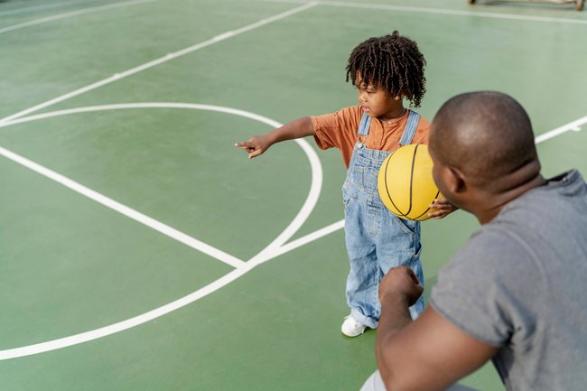 Ein schwarzes Kind hat einen gelben Basketball in der Hand und zeigt auf den Sportplatz.