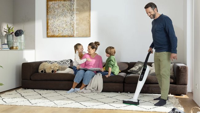 In einem Wohnzimmer saugt der Mann staub, während die mutter mit den Kindern auf der Couch sitzt und ein Buch vorliest.