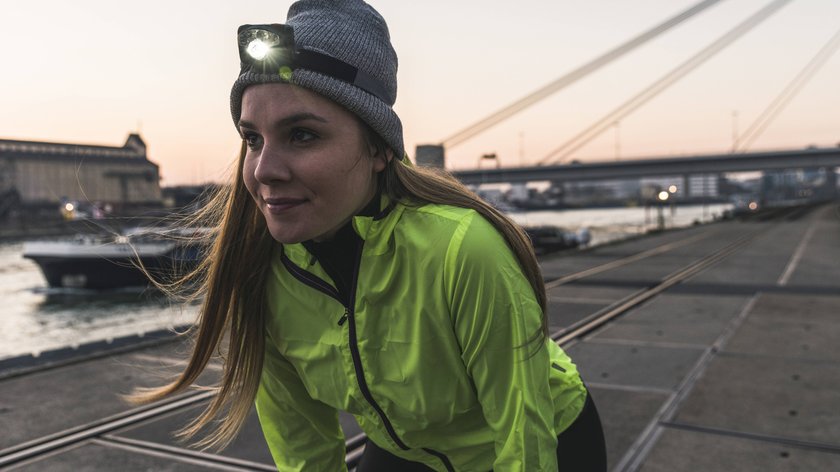 Eine Joggerin in neon-gelber Jacke mit Stirnlampe. Im Hintergrund ist ein Hafen.