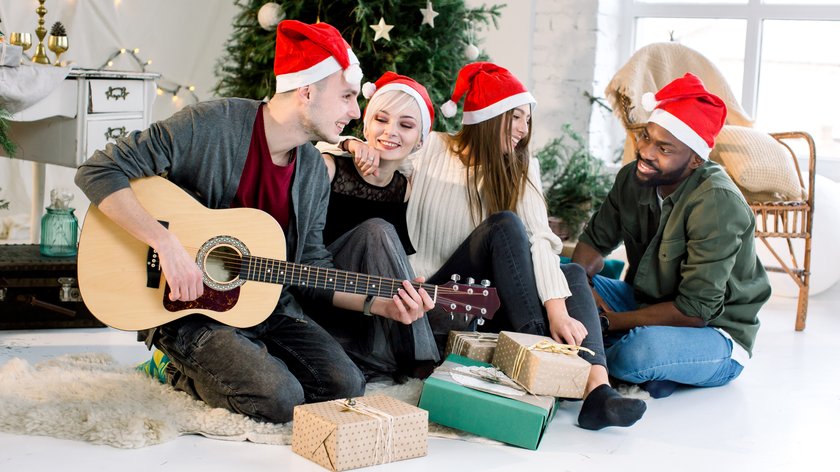 Picture showing group of four friends celebrating Christmas at home. Young Caucasian man is playing guitar and the girls and African man are smiling and singing carols