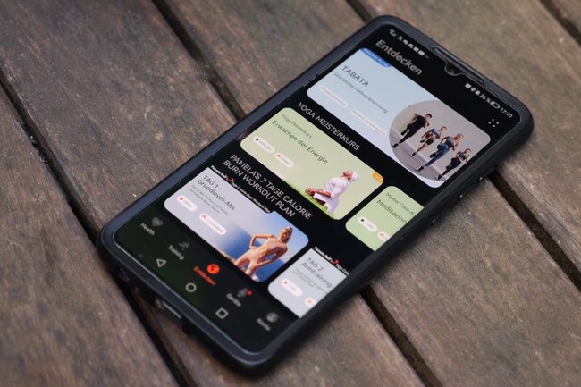 Ein Smartphone liegt auf einem Holzuntergrund. Auf dem Display sieht man die Hauwei Health App mit den vielen Fitnessangeboten.